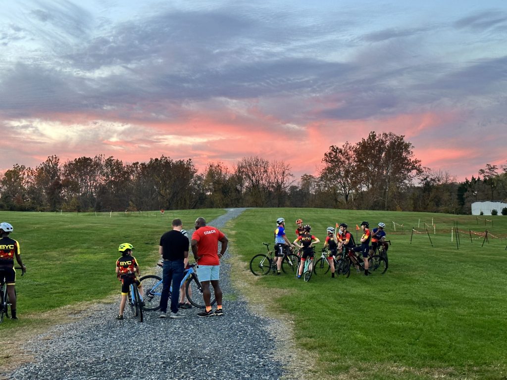 Photo of a group of adults looking at a bike on a gravel path. To the side is a group of kids on bikes gathered on a grassy field.