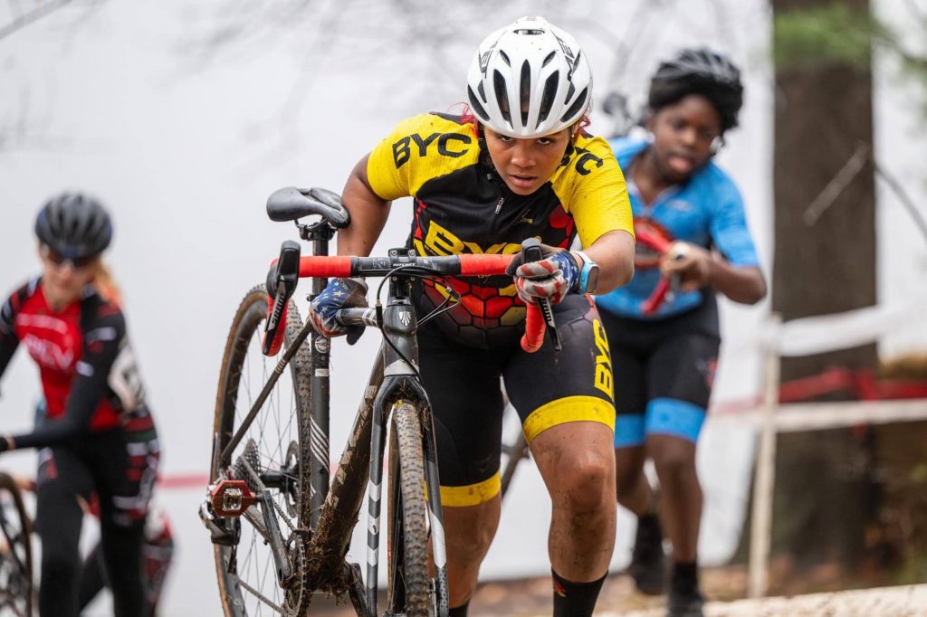 Photograph of a biker in BYC kit rolling a muddy bike with an expression of focus on their face