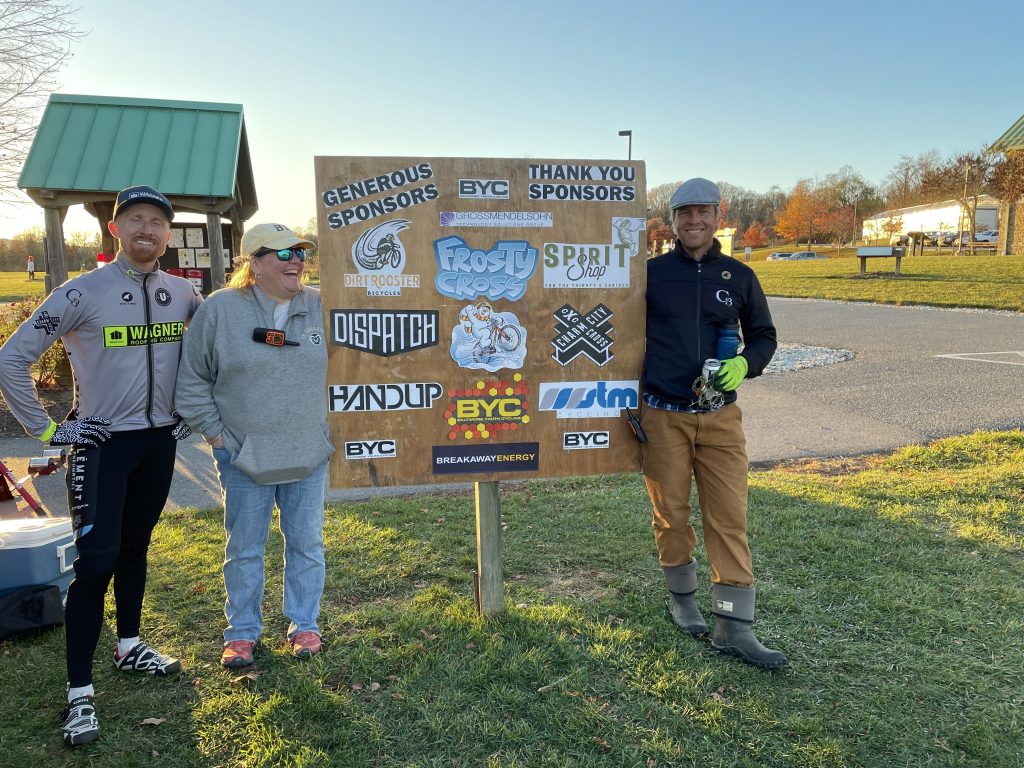 Photograph of three adults standing beside board listing sponsors of Frosty Cross including Dirt Rooster, Gross Mendelsohn, Spirit Shop, Dispatch, Charm City Cross, Handup, STM, and Breakaway Energy