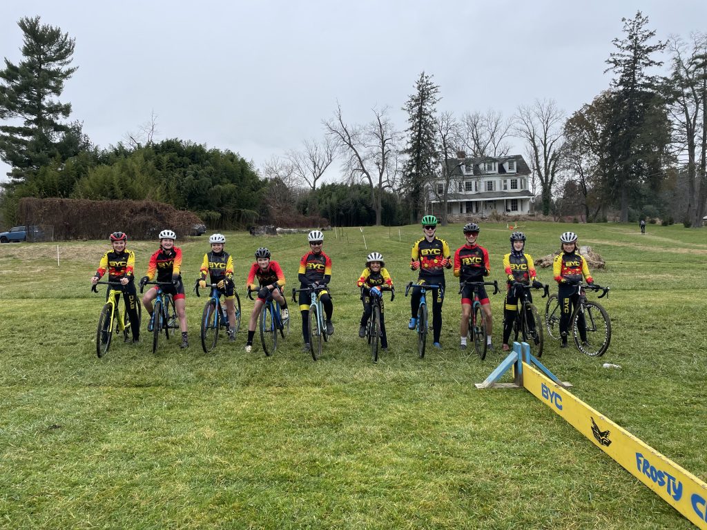 Photo of ten kids standing with bikes behind a cyclocross barrier